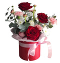 Arreglo de rosas rojas, ranúnculos y margaritas blancas en una caja redonda roja con lazo rosa